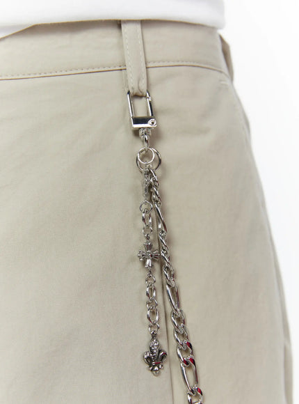 Men's Layered Cross Chain IL509 Silver close-up detail on beige trousers showing carabiner clip cross charm and fleur-de-lis charm with layered figaro chain