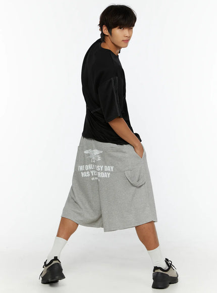 Men's Lettering Bermuda Shorts IL513 Gray three-quarter back view on male model in motion, back graphic print visible, black tee, hands in pockets, sneakers and white socks