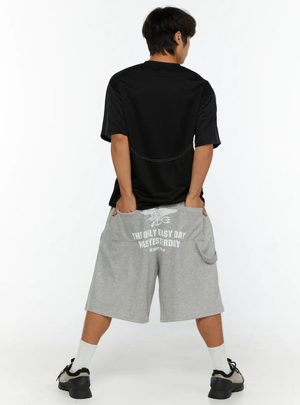 Men's Lettering Bermuda Shorts IL513 Gray back view on male model, white US Navy Seals eagle graphic print and lettering across seat, hands in pockets, black tee styling
