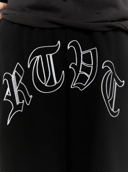 Men's Logo Embroidered Snap Hem Bermuda Sweat Shorts (Black) IL507 close-up detail of white gothic lettering arc embroidery on black fabric showing stitch texture