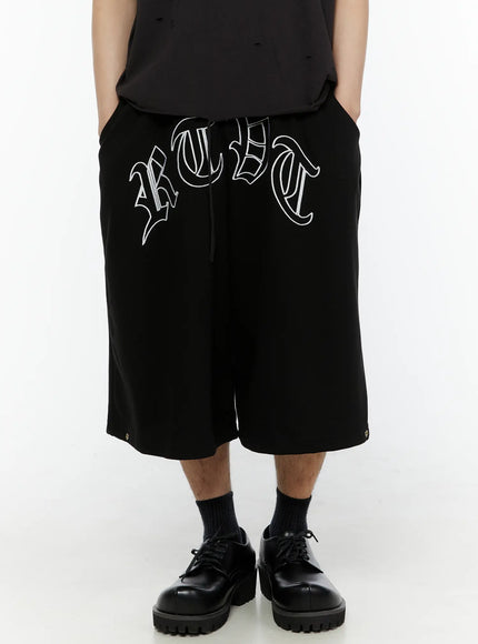 LEWKIN Men's Logo Embroidered Snap Hem Bermuda Sweat Shorts (Black) IL507 front view on model hands in pockets paired with black distressed tee and derby shoes