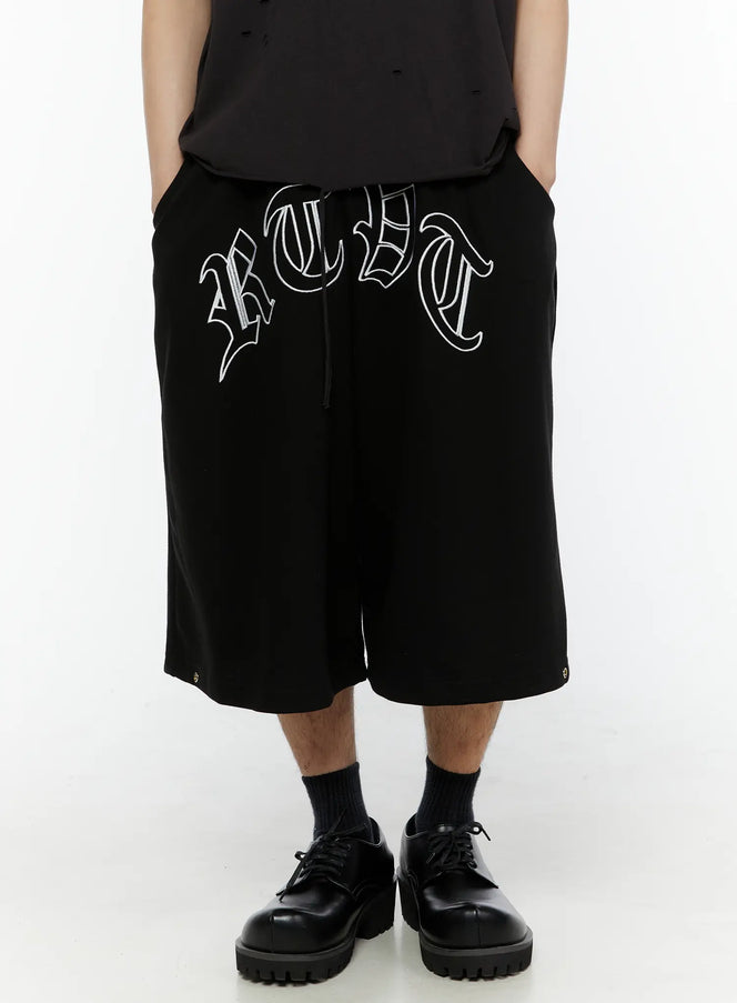LEWKIN Men's Logo Embroidered Snap Hem Bermuda Sweat Shorts (Black) IL507 front view on model hands in pockets paired with black distressed tee and derby shoes