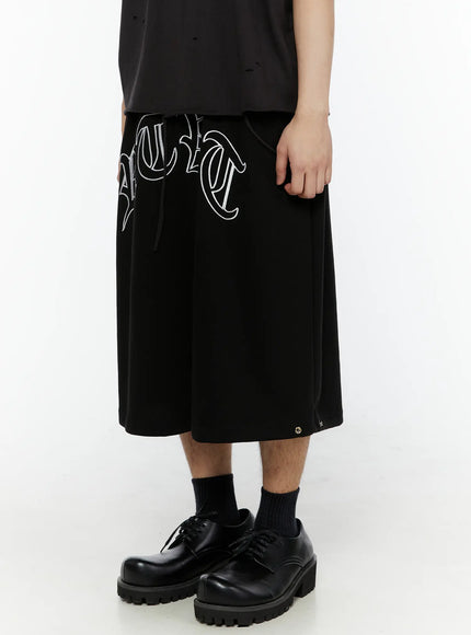 Men's Logo Embroidered Snap Hem Bermuda Sweat Shorts (Black) IL507 side view on model showing gothic arc logo embroidery and wide-leg silhouette with black derby shoes