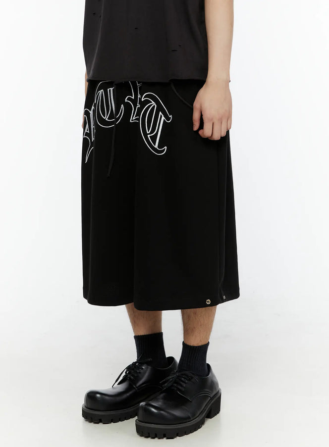 Men's Logo Embroidered Snap Hem Bermuda Sweat Shorts (Black) IL507 side view on model showing gothic arc logo embroidery and wide-leg silhouette with black derby shoes