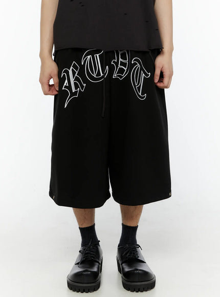 Men's Logo Embroidered Snap Hem Bermuda Sweat Shorts (Black) IL507 front close-up on model highlighting oversized gothic arch logo embroidery with white outline stitching