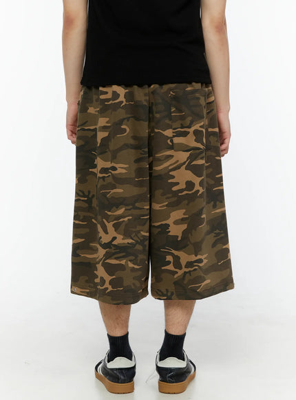 Men's Logo Embroidered Snap Hem Bermuda Sweat Shorts (Camouflage) IL507 Camouflage three-quarter back view on model carrying black leather bag, old-English embroidery visible on right thigh, wide-leg bermuda cut