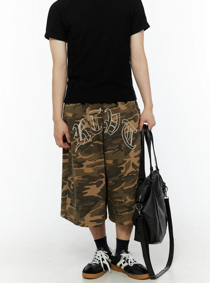 Men's Logo Embroidered Snap Hem Bermuda Sweat Shorts (Camouflage) IL507 Camouflage front view on model with black tee, hands holding hem, elastic waistband and large old-English RTBT embroidery prominent