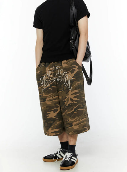 Men's Logo Embroidered Snap Hem Bermuda Sweat Shorts (Camouflage) IL507 Camouflage three-quarter front view on model with black tee and leather crossbody bag, embroidery and wide-leg silhouette shown from slight angle