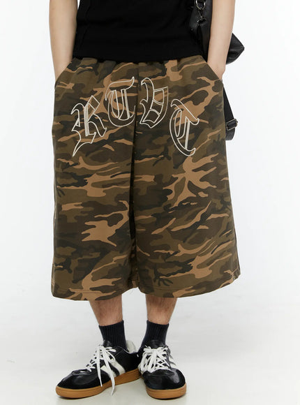 LEWKIN Men's Logo Embroidered Snap Hem Bermuda Sweat Shorts (Camouflage) IL507 Camouflage front view on model wearing black tee and sneakers, hands in pockets, oversized wide-leg silhouette with old-English embroidery