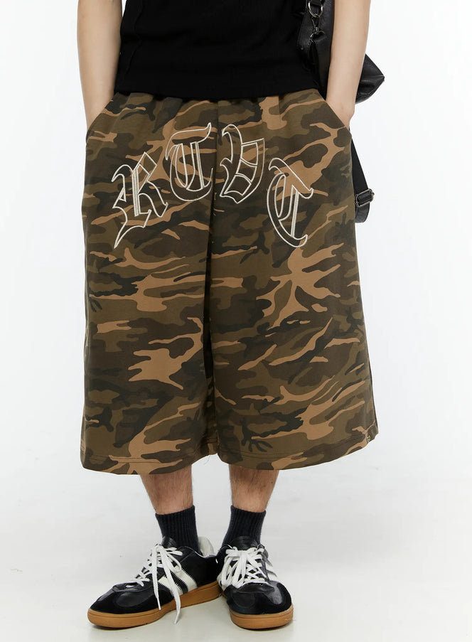 LEWKIN Men's Logo Embroidered Snap Hem Bermuda Sweat Shorts (Camouflage) IL507 Camouflage front view on model wearing black tee and sneakers, hands in pockets, oversized wide-leg silhouette with old-English embroidery