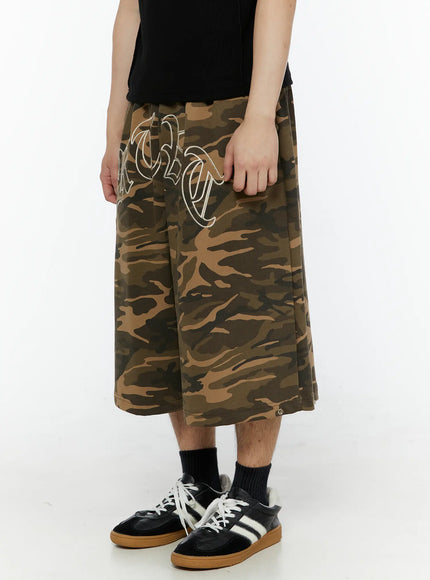 Men's Logo Embroidered Snap Hem Bermuda Sweat Shorts (Camouflage) IL507 Camouflage side-angled view on model, left hand gripping fabric, old-English embroidered lettering visible, wide-leg cotton bermuda length