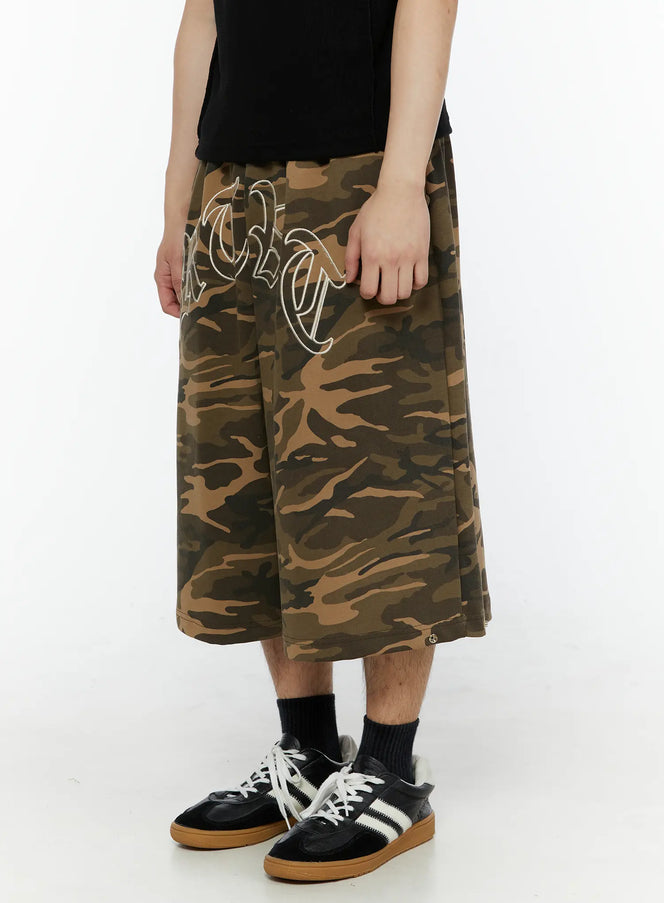Men's Logo Embroidered Snap Hem Bermuda Sweat Shorts (Camouflage) IL507 Camouflage side-angled view on model, left hand gripping fabric, old-English embroidered lettering visible, wide-leg cotton bermuda length