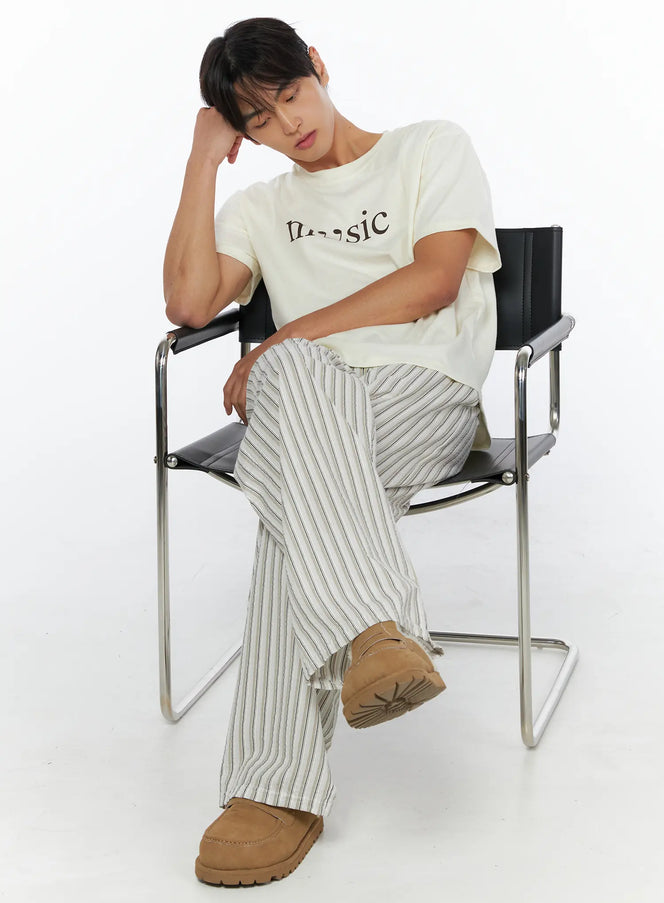 LEWKIN Men's Music Print T-Shirt IL515 ivory seated lifestyle shot male model resting head on hand on black metal chair wearing striped wide-leg pants