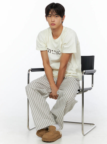 Men's Music Print T-Shirt IL515 ivory front seated shot male model leaning forward hands clasped paired with striped wide-leg trousers and tan mules