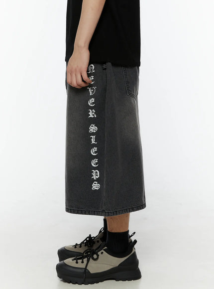 Men's 'Never Stop' Graphic Bermuda Jorts (Dark Gray) IL507 side view on model showing full-length right leg vertical old English letter graphic on washed dark gray denim