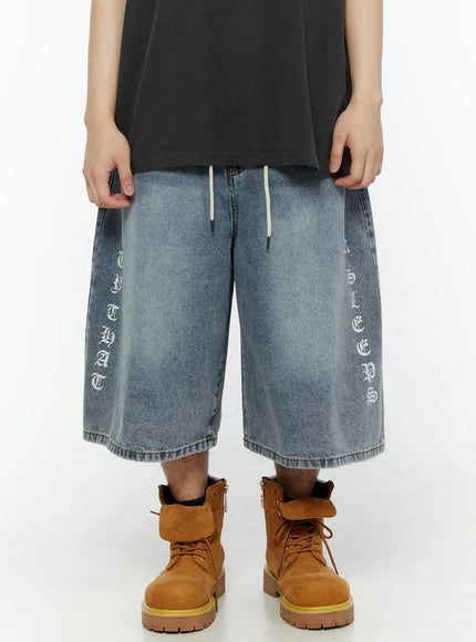 LEWKIN Men's 'Never Stop' Graphic Bermuda Jorts (Light Blue) IL507 front view on model, washed light blue denim with old-english vertical letter prints on both legs, cream drawstring waist, styled with tan boots