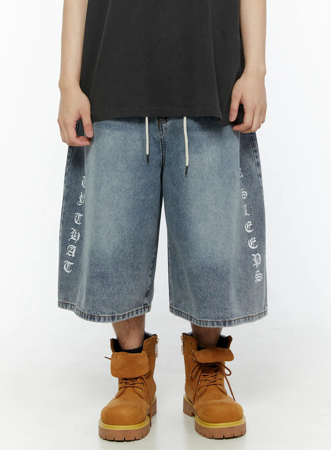 LEWKIN Men's 'Never Stop' Graphic Bermuda Jorts (Light Blue) IL507 front view on model, washed light blue denim with old-english vertical letter prints on both legs, cream drawstring waist, styled with tan boots