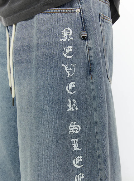 Men's 'Never Stop' Graphic Bermuda Jorts (Light Blue) IL507 close-up detail of old-english vertical graphic lettering on right leg side panel, washed light blue denim texture visible