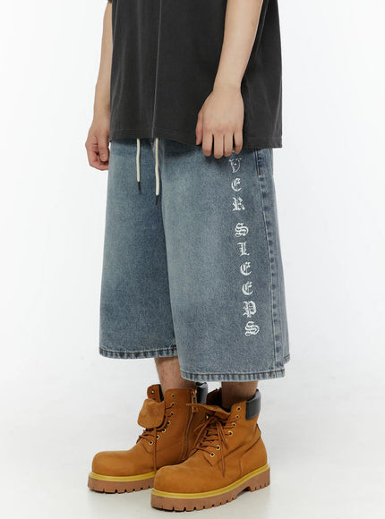 Men's 'Never Stop' Graphic Bermuda Jorts (Light Blue) IL507 left side close-up on model, wide-leg denim with bold old-english letter graphic print running vertically, cream drawstring detail at waist