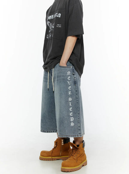 Men's 'Never Stop' Graphic Bermuda Jorts (Light Blue) IL507 side view on model, right leg showing full 'NEVER SLEEPS' old-english vertical graphic print, wide-leg silhouette, styled with graphic tee and tan boots