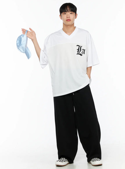 Men's Oversized Mesh Rugby Tee IL514 White front full-body shot on male model holding light blue cap in raised hand, black La gothic logo on chest, oversized V-neck silhouette