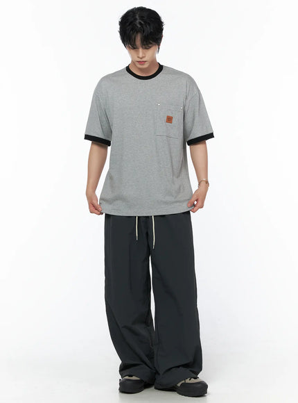 Men's Patch Detail Short Sleeve Tee IU523 Gray front view on male model looking down, black ringer trim, chest pocket with leather patch, styled with dark wide-leg pants