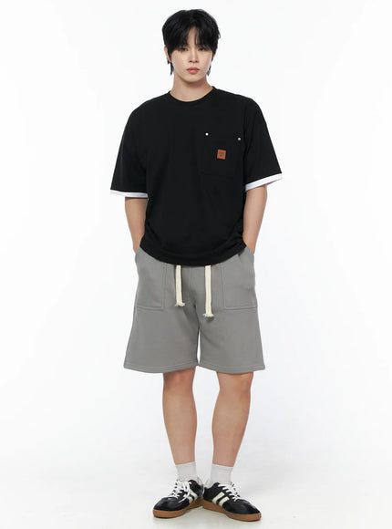 Men's Patch Detail Short Sleeve Tee IU523 Black front view on male model standing with hands in pockets, white ringer trim, chest pocket with leather patch, styled with gray drawstring shorts