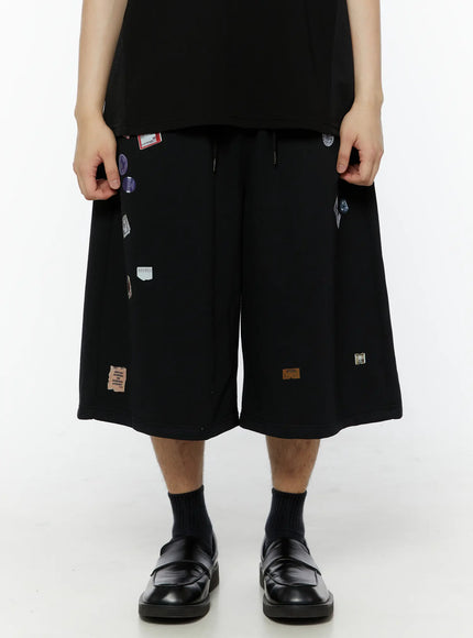 LEWKIN Men's Patch Detail Shorts (Black) IL509 black front close-up on model, wide-leg cut with scattered colorful sticker patches, drawstring waist, styled with loafers