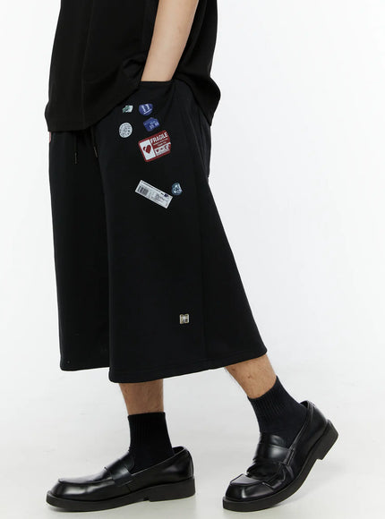 Men's Patch Detail Shorts (Black) IL509 black side close-up on model, hand in pocket, showing Fragile label and barcode sticker patches on wide-leg silhouette