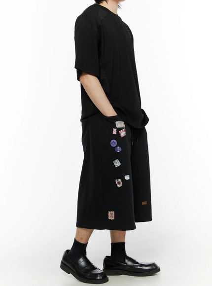 Men's Patch Detail Shorts (Black) IL509 black side full-length on model, oversized T-shirt styling, wide-leg shorts with scattered patch prints and loafers