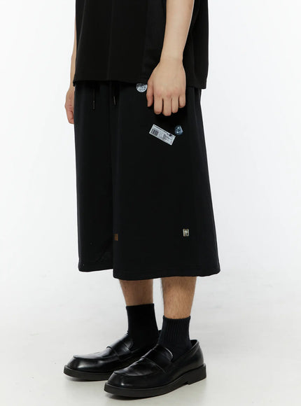 Men's Patch Detail Shorts (Black) IL509 black side close-up on model, hand gripping fabric, barcode and sticker patches visible, wide-leg shorts with drawstring waist