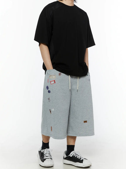 LEWKIN Men's Patch Detail Shorts (Gray) IL509 gray angled side view on model styled with black oversized tee and sneakers showing scattered label patches