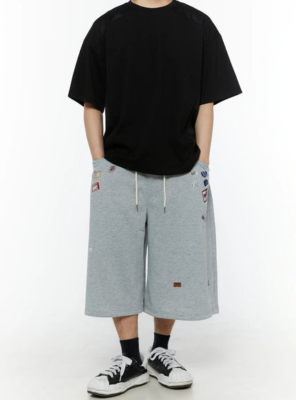 Men's Patch Detail Shorts (Gray) IL509 gray front full-body shot on male model hands in pockets showing all-over label patch graphics and ivory drawstring waist