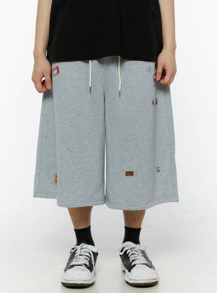 Men's Patch Detail Shorts (Gray) IL509 gray front close-up on model hands gripping waistband showing wide-leg cut drawstring and scattered patch graphics