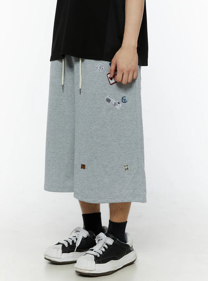 Men's Patch Detail Shorts (Gray) IL509 gray side view on male model right hand holding red patch card showing wide-leg hem and label badge graphics