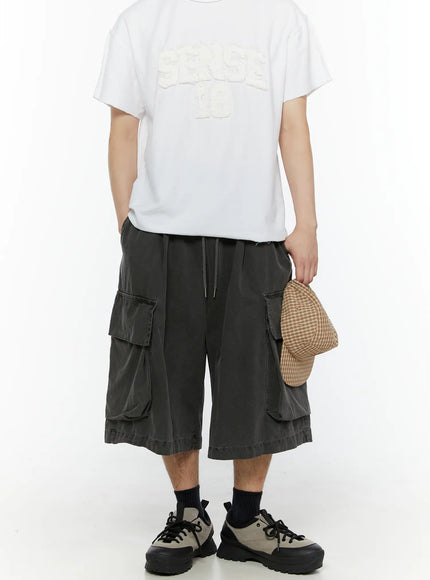 LEWKIN Men's Pigment Bermuda Cargo Shorts (Dark Gray) IL507 Dark gray front model shot full-length styled with white graphic tee and sneakers holding plaid cap