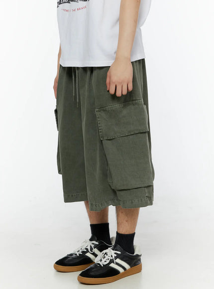Men's Pigment Bermuda Cargo Shorts (Dark Green) IL507 Dark green left side view on male model hand resting on cargo pocket showing layered hem and washed texture