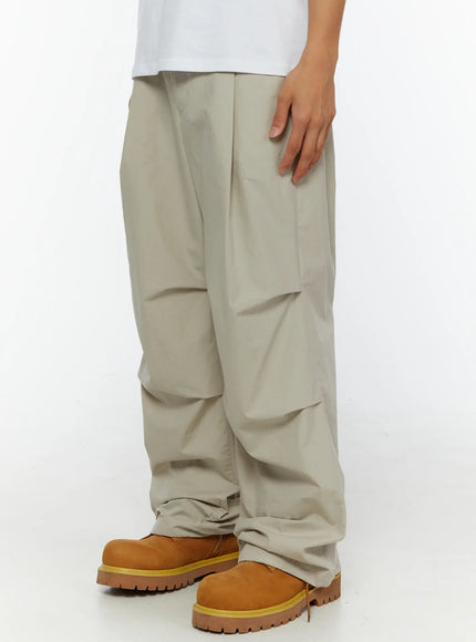 Men's Relaxed-Fit Parachute Pants IL509 Light beige angled front view on model showing wide-leg drape and crinkled nylon texture with tan boots