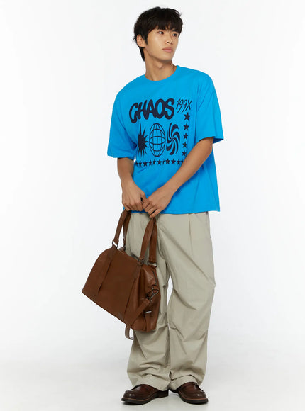Men's Relaxed-Fit Parachute Pants IL509 Light beige three-quarter front shot on model in blue graphic tee holding brown bag looking aside with loafers