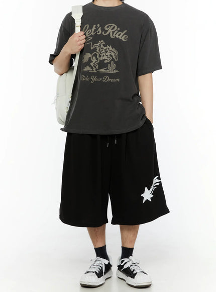 LEWKIN Men's Shooting Star Printed Shorts (Black) IL509 front view on male model styled with grey graphic tee and white backpack, wide-leg silhouette with white shooting star print