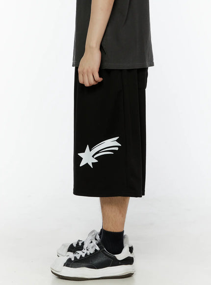 Men's Shooting Star Printed Shorts (Black) IL509 side close-up on male model showing white shooting star print placement on lower left leg panel and wide-leg silhouette