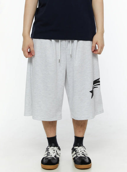 Men's Shooting Star Printed Shorts (Light Gray) IL509 Light gray front view on model holding waistband, white drawstring detail, shooting star print at hem, wide-leg cut