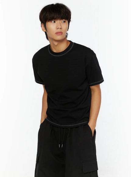 Men's Stitched Cropped T-Shirt IL513 Black upper-body front shot on male model looking away, hands in pockets, contrast stitching visible at neckline and hem