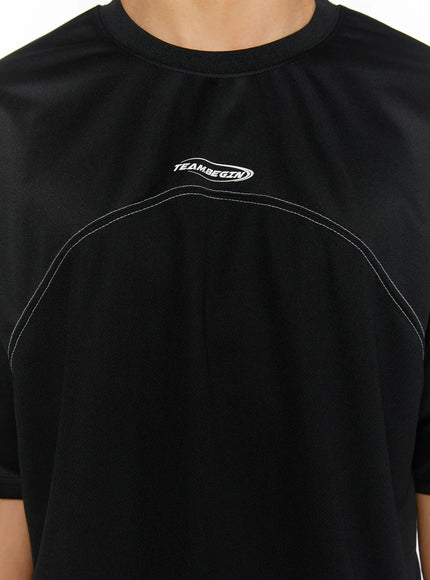 Men's Stitched T-Shirt IL513 Black close-up front detail showing TEAM.BEGIN oval logo and double white contrast stitch arch on chest