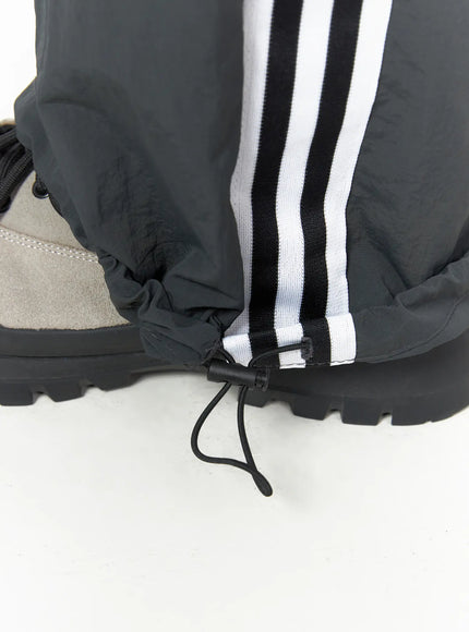 Men's Striped Wide Track Pants IU523 dark gray close-up detail of ankle hem showing adjustable drawcord toggle and white side stripe tape on nylon fabric