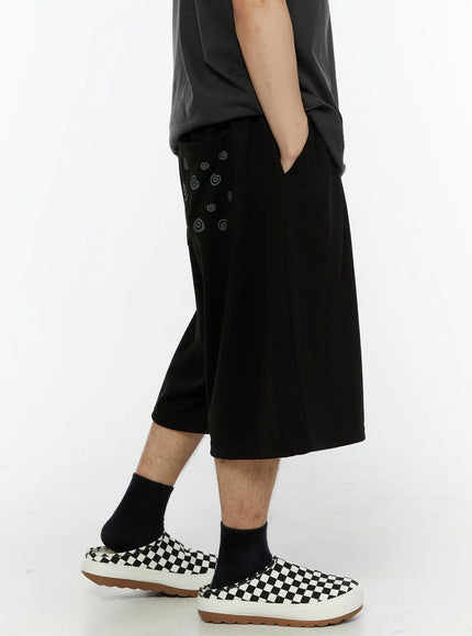 Men's Swirl Print Back Pocket Shorts (Black) IL509 Black side shot on model showing swirl graphic pocket detail wide-leg cut styled with graphic sleeveless tee and checkered slip-ons