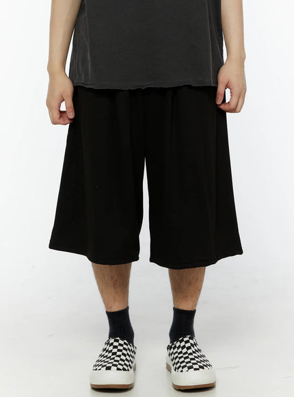 Men's Swirl Print Back Pocket Shorts (Black) IL509 Black front close-up shot on model showing wide-leg silhouette elastic waistband and relaxed fit with checkered slip-on shoes