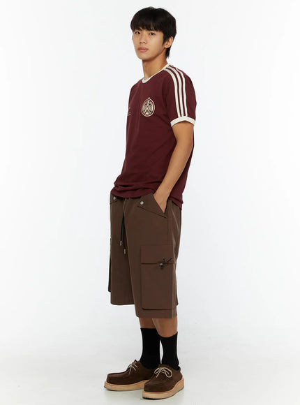 Men's Track Graphic T-Shirt IL507 Burgundy three-quarter side on model, hands in pockets, styled with brown cargo shorts, black socks and suede shoes