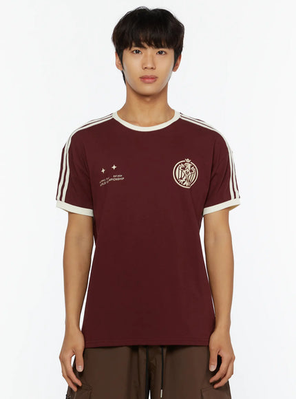 Men's Track Graphic T-Shirt IL507 Burgundy cropped front close-up on model, cream crew neck collar, shoulder stripes, eagle crest and star graphic clearly visible