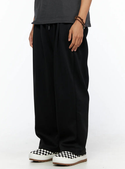 Men's Triple Tuck Sweatpants IL520 Black angled side view on model showing wide-leg drape, pleated front, and drawstring elastic waistband with checkered slip-on sneakers